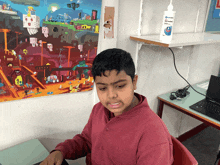 a boy sits at a desk with a laptop and a bottle of hand sanitizer on a shelf behind him