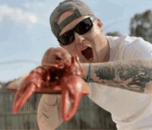 a man wearing sunglasses and a baseball cap is holding a lobster in his hands .