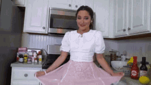 a woman in a white top and pink skirt is standing in a kitchen with bottles of hot sauce on the counter