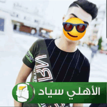 a man wearing sunglasses and a smiley face on his face stands in front of a green sign with arabic writing