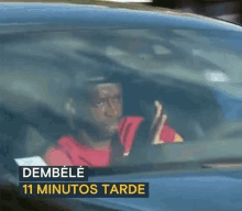 a man in a red shirt is driving a blue car with the words dembele 11 minutos tarde written on the bottom .