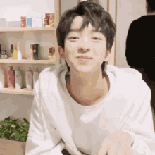 a young man wearing a white shirt is sitting in front of a shelf with bottles of cosmetics on it .