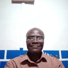 a man wearing glasses and a brown shirt is sitting in front of a cross .