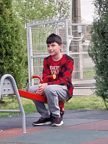 a boy wearing a red plaid shirt that says den visi sits on a red seesaw