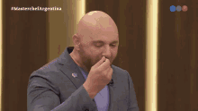 a bald man in a suit is covering his nose in front of a screen that says #masterchefargentina