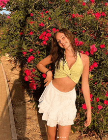 a woman in a white skirt and a green crop top is standing in front of a bush with pink flowers