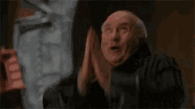 a bald man in a black coat is making a funny face while standing in front of a window .