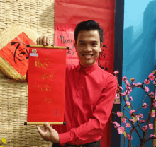 a man in a red shirt is holding a scroll with chinese writing on it in front of a bamboo wall