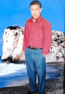 a young man in a red shirt and blue pants is standing on a beach in front of a large rock .