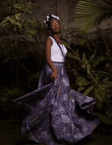 a woman wearing a white tank top and a blue skirt is standing in the woods