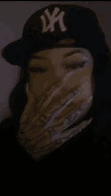 a woman with long nails is wearing a ny hat and covering her face with her hand .