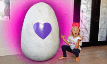 a little girl with a surprised look on her face is kneeling next to a large egg with a heart on it