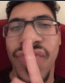 a man wearing glasses and a beard is holding his finger to his nose .