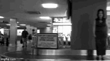 a black and white photo of people walking around a conveyor belt with a sign that says keep off conveyor belts