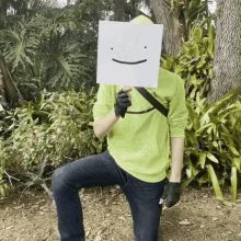 a man in a yellow shirt is holding a paper with a smiley face on it in front of his face