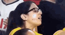 a woman wearing glasses and a yellow shirt is sitting in a stadium with a sticker on her face .