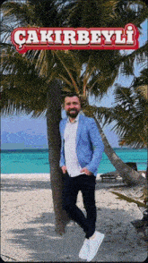 a man in a blue jacket leans against a palm tree on a beach with the name çakirbeyli above him