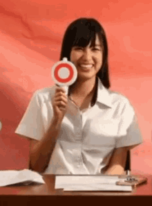 a woman in a white shirt is sitting at a table holding a sign with a red circle on it .