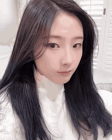 a woman with long black hair wearing a white turtleneck looks at the camera