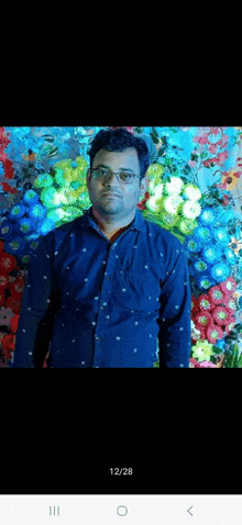 a man wearing glasses is standing in front of a wall of flowers .