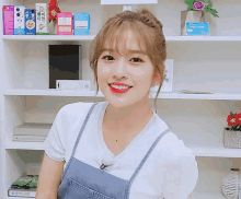 a woman in a white shirt and blue overalls smiles in front of a shelf with boxes of food on it