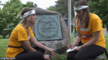 two girls in yellow shirts are kneeling in front of a stone with a plaque on it that says fair valley school