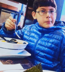 a young boy in a blue jacket is eating a bowl of soup with a fork