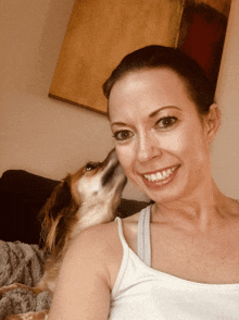 a woman in a white tank top is smiling while a dog kisses her on the cheek