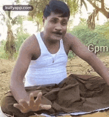 a man in a white tank top and brown pants is sitting on the ground with his hands outstretched .