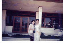 two women pose for a picture in front of a building with a sign that says dilarang masuk