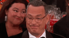 a man wearing glasses and a tuxedo is making a funny face while a woman looks on .