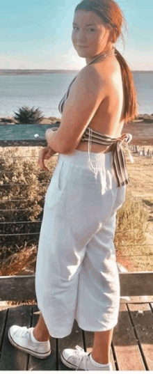 a woman in a bikini top and white pants is standing on a wooden deck overlooking the ocean .