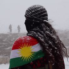 a woman with dreadlocks is holding a kurdish flag on her back in the snow