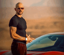 a bald man wearing sunglasses and a black shirt is standing next to a red sports car