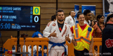 a man with a blue belt stands in front of a scoreboard that says gustavo santos and peter leung