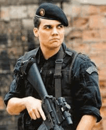 a man in a military uniform is holding a gun in front of a brick wall .