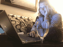 a woman sits on a couch using a laptop and a glass of wine