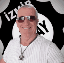 a man wearing sunglasses and a necklace smiles in front of an izmir ay logo