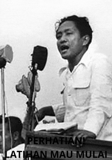 a black and white photo of a man speaking into a microphone with the words perhatiani latihan mau mulai on the bottom