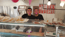 two men behind a counter with a menu on the wall behind them that says ' sandwiches ' on it