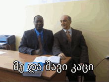 two men are sitting at a desk with a foreign language written on the table