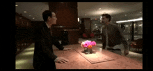 a man in a suit is standing at a counter with a vase of pink flowers on it