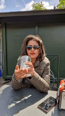 a woman wearing sunglasses sits at a table holding a glass of water