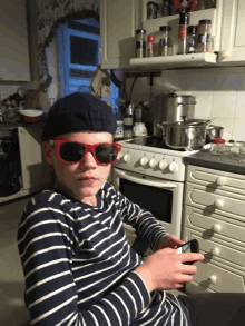 a boy wearing sunglasses and a beret sits in front of a stove