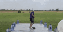 a man in a purple shirt is standing on a white tarp in a field with water bottles in the background .