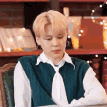 a young man wearing a green vest and tie is sitting at a table in front of a bookshelf .