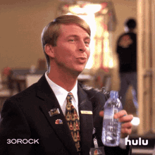 a man in a suit and tie is holding a bottle of water with the hulu logo in the background