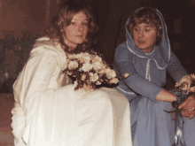 a woman in a white dress sits next to a woman in a blue dress holding a bouquet of flowers