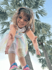 a little girl wearing a rainbow dress and pink shoes reaches out her hands