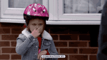 a little girl wearing a pink helmet holds her nose in front of a brick wall that says coronation st.